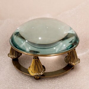 May include: A large, clear magnifying glass with a teal tint, set on a brass base. The base features a circular design supported by three ornate, cone-shaped legs. The magnifying glass is designed for reading or detailed work.