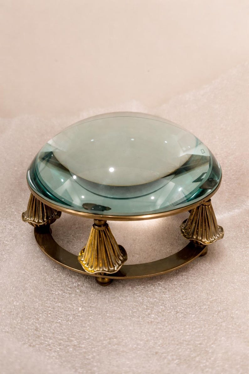 May include: A large, clear magnifying glass with a teal tint, set on a brass base. The base features a circular design supported by three ornate, cone-shaped legs. The magnifying glass is designed for reading or detailed work.