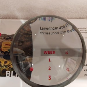 May include: A circular magnifying glass with a dark metal rim, partially obscuring a white paper with text and numbers. The text reads "Leave those thrives under the he" and "WEEK 1 2 3 4". A DHL truck is visible in the background.