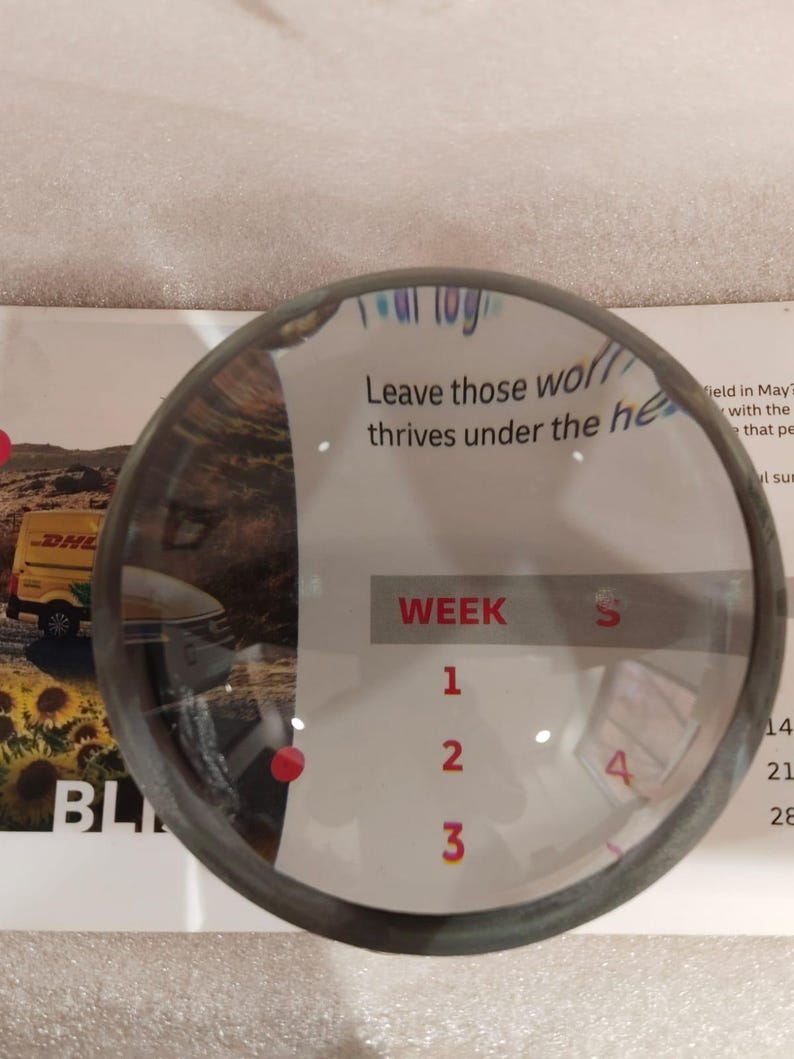 May include: A circular magnifying glass with a dark metal rim, partially obscuring a white paper with text and numbers. The text reads "Leave those thrives under the he" and "WEEK 1 2 3 4". A DHL truck is visible in the background.