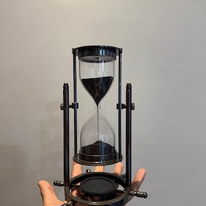 Op de afbeelding: Zandloper met zwart zand, in een donker metalen frame. Het frame heeft een cirkelvormige basis met decoratieve spaken. De glazen bol is helder en het zand is geconcentreerd in de onderste helft.