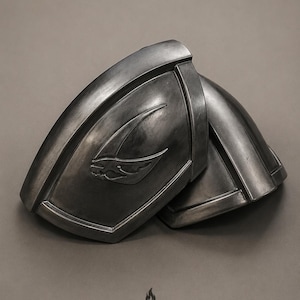 May include: A pair of dark metallic shoulder pauldrons featuring a curved, molded design with a raised, stylized emblem on the front surface. The armor pieces have a weathered, gunmetal finish with defined edges and a smooth, contoured shape.
