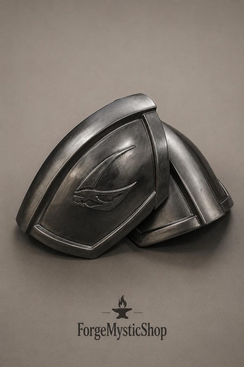 May include: A pair of dark metallic shoulder pauldrons featuring a curved, molded design with a raised, stylized emblem on the front surface. The armor pieces have a weathered, gunmetal finish with defined edges and a smooth, contoured shape.