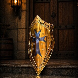 Op de afbeelding: Een goudkleurig schild met een blauw kruisontwerp. Het schild heeft een decoratieve rand en staat tegen een donkere, getextureerde achtergrond, wat een middeleeuws thema suggereert. Het schild is ongeveer 61 cm hoog.