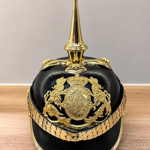 Op de afbeelding: Een zwarte helm met gouden accenten, waaronder een opvallende piek aan de bovenkant. De helm heeft een gouden wapen met twee leeuwen en een kroon. Een gouden, kettingachtige rand loopt langs de basis van de helm.