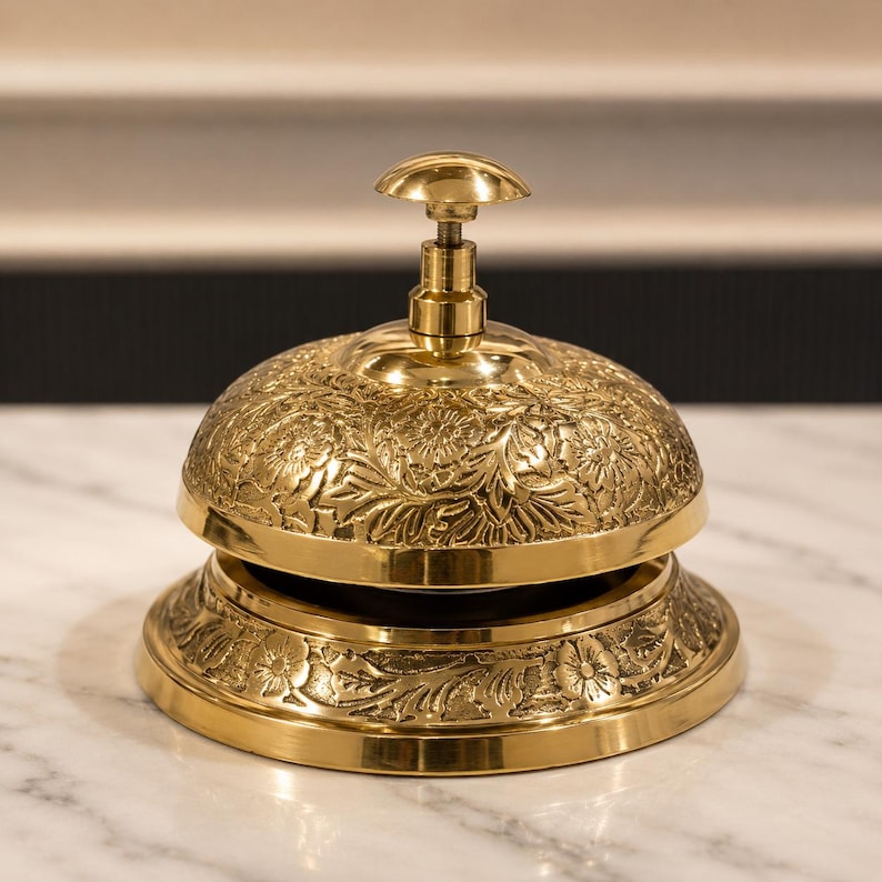 Peut inclure: Une cloche de r&eacute;ception en laiton poli pr&eacute;sentant un motif floral et v&eacute;g&eacute;tal en relief complexe sur le d&ocirc;me et la base circulaire. La cloche est dot&eacute;e d'un bouton-poussoir vertical central au sommet, con&ccedil;u pour une utilisation manuelle.