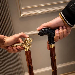 Op de afbeelding: Twee wandelstokken met houten schachten en decoratieve handvatten. Eén handvat is een gepolijste goudkleurige leeuwenfiguur en de andere is een matzwarte dierenkopfiguur. Beide schachten hebben een donkerbruine houtnerf met kleine accenten.