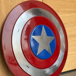 K&ouml;nnte beinhalten: Runder Schild im Captain America-Stil mit einem roten Au&szlig;enring, einem silbernen Innenring und einer blauen Mitte mit einem silbernen Stern. Der Schild ist &uuml;berwiegend rot, silber und blau.