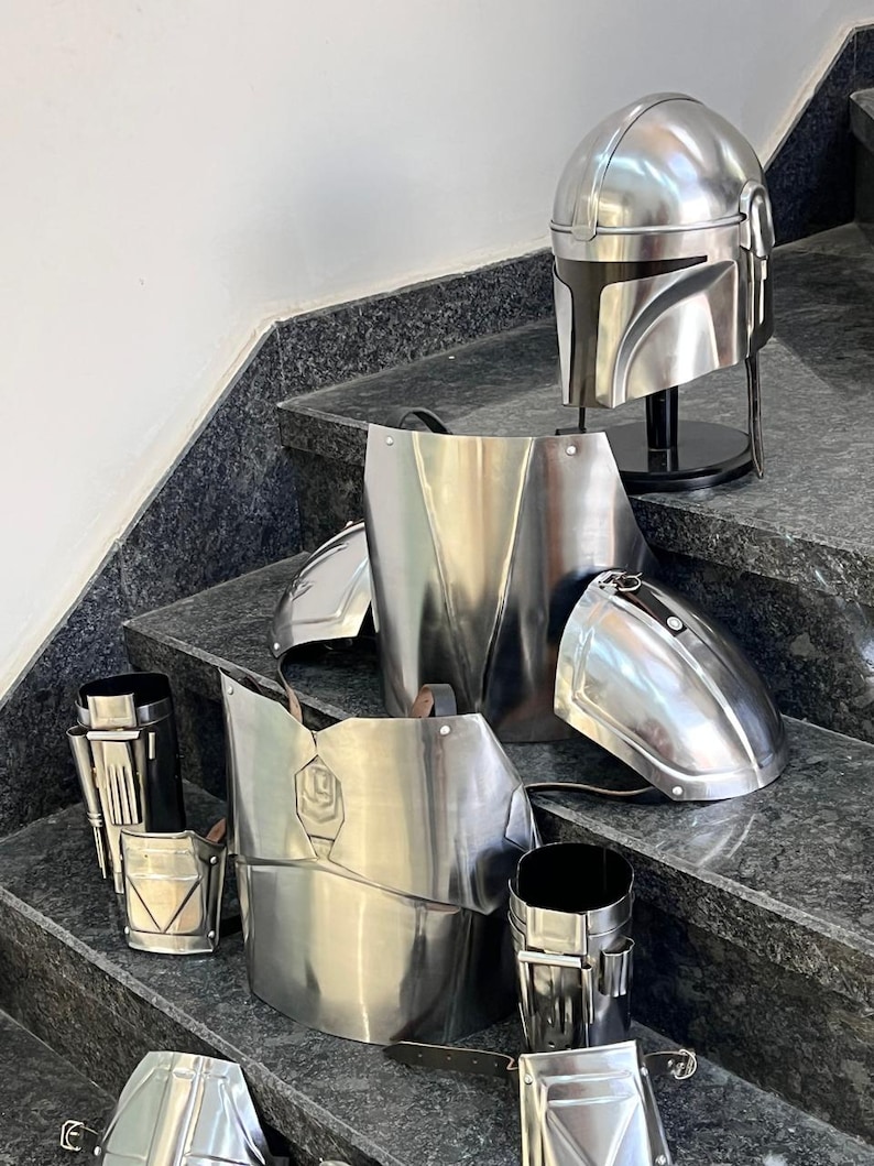 May include: A complete set of shiny, silver-colored Mandalorian armor, including a helmet, chest plate, shoulder pads, and other components. The armor is displayed on a set of stairs, showcasing its intricate design and metallic finish.