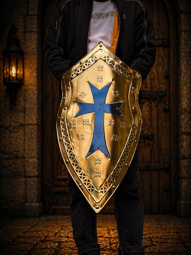 Op de afbeelding: Een goudkleurig schild met een blauw kruisontwerp. Het schild heeft een decoratieve rand en wordt door een persoon vastgehouden. De achtergrond toont een houten deur en een lantaarn.