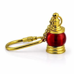 May include: A miniature red and gold lantern keychain. The lantern has a round, red glass body with gold-colored metal accents, including a chain and a key ring. The keychain is designed to resemble a traditional lantern.