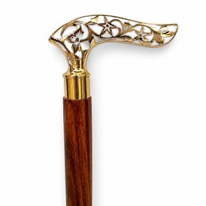 May include: A walking cane featuring a dark brown wooden shaft and a polished gold-tone handle. The handle is shaped in a curved design and is decorated with white enamel inlays and intricate gold floral patterns throughout the surface.