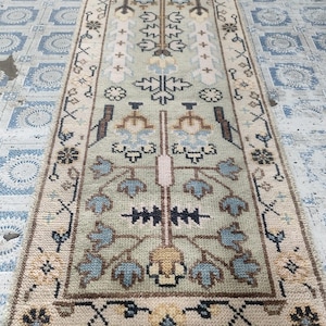 May include: A rectangular, hand-knotted rug with a central design in shades of green, beige, and blue. The rug features floral and geometric patterns, with a cream-colored border and fringe. The rug's dimensions are approximately 2 x 4 feet.