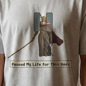 May include: A light grey t-shirt with a graphic of a character with a staff and cape, framed by two vertical rectangles. The text "Paused My Life for This Boss" is printed below the graphic in a white rectangle.