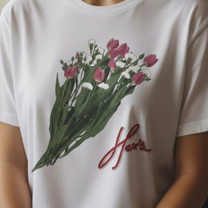 May include: White t-shirt featuring a bouquet of pink tulips and white flowers with green stems. The word "Her's" is written in red script below the flowers. The shirt is a classic crew neck style.