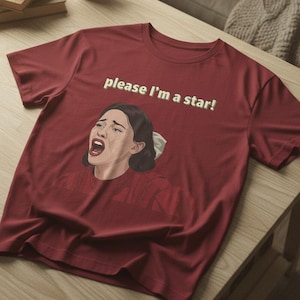 Może przedstawiać: Bordowa koszulka z napisem "please I'm a star!" i grafiką kobiety. Koszulka leży na drewnianym stole.