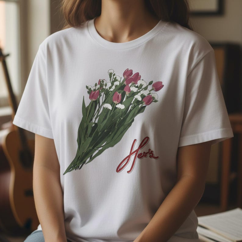May include: White t-shirt featuring a bouquet of pink tulips and white flowers with green stems. The word "Her's" is written in red script below the flowers. The shirt is a classic crew neck style.