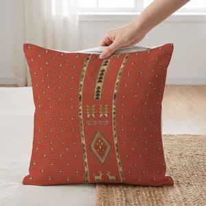 May include: A square throw pillow in a burnt orange hue featuring a central vertical band with geometric patterns, including triangles, diamonds, and two small animal figures. The surrounding areas are covered in a repeating small triangle motif.