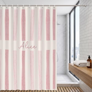 May include: A shower curtain featuring a pattern of vertical light pink watercolor-style stripes on a cream background. The name Alice is printed in a cursive pink script font across the center of the curtain, which hangs from a metal rod.