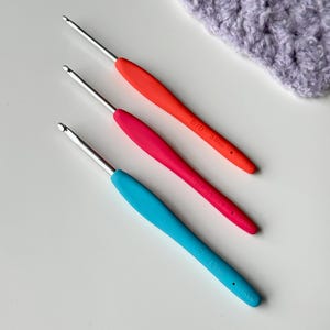 May include: Three crochet hooks with colorful handles in red, pink, and blue. The hooks are made of metal and have measurements of 3.0mm, 3.5mm, and 4.5mm. A piece of lavender crochet fabric is in the background.