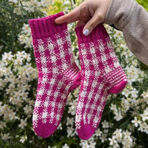 May include: A pair of mid-calf socks featuring a vibrant magenta and white checkered pattern. The socks have a ribbed cuff at the top and a solid magenta toe and heel section, constructed with a knit texture throughout the fabric.