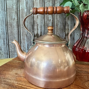 May include: A copper tea kettle with a wooden handle and lid knob. The kettle has a spout and a curved handle. The copper has a shiny finish. The kettle is sitting on a wooden surface.