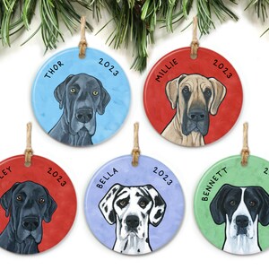 May include: Five round ceramic Christmas ornaments featuring Great Dane dog portraits. Each ornament has a different colored background and a dog's name and the year 2023 printed on it. The names are Thor, Millie, Bailey, Bella, and Bennett.
