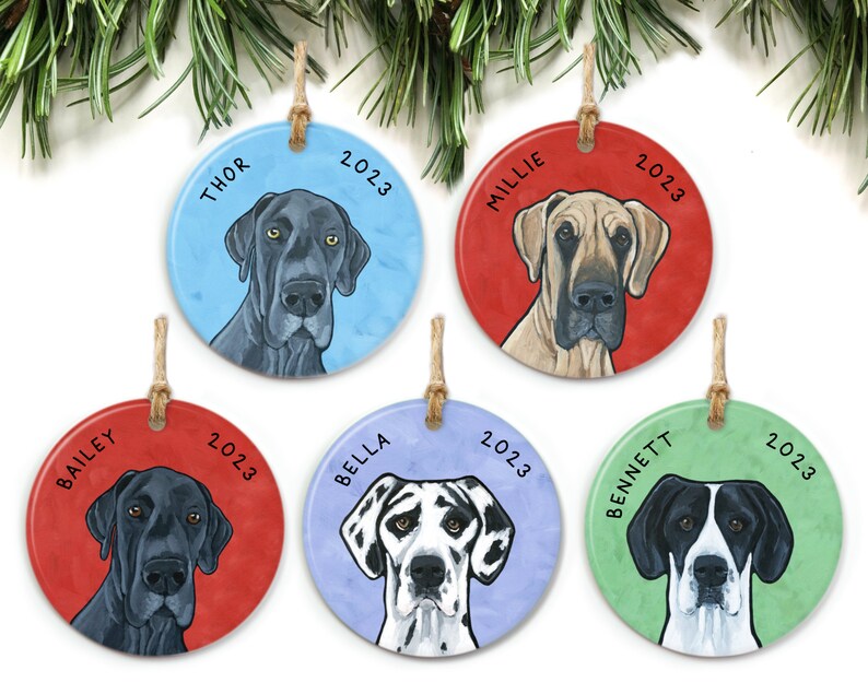 May include: Five round ceramic Christmas ornaments featuring Great Dane dog portraits. Each ornament has a different colored background and a dog's name and the year 2023 printed on it. The names are Thor, Millie, Bailey, Bella, and Bennett.