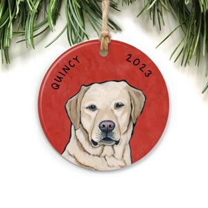 Puede incluir: Un adorno navide&ntilde;o redondo y rojo con una ilustraci&oacute;n de un perro Labrador Retriever blanco. El nombre del perro, "QUINCY", est&aacute; escrito en el lado izquierdo del adorno, y el a&ntilde;o "2023" est&aacute; escrito en el lado derecho.