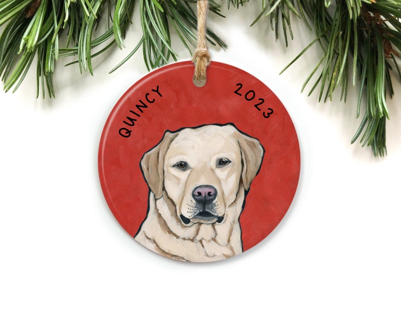 Puede incluir: Un adorno navide&ntilde;o redondo y rojo con una ilustraci&oacute;n de un perro Labrador Retriever blanco. El nombre del perro, "QUINCY", est&aacute; escrito en el lado izquierdo del adorno, y el a&ntilde;o "2023" est&aacute; escrito en el lado derecho.