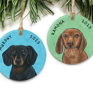 May include: Two round Christmas ornaments with dachshund dog portraits. The ornament on the left is blue with the name "MURPHY" and the year "2023". The ornament on the right is green with the name "RAMONA" and the year "2023".