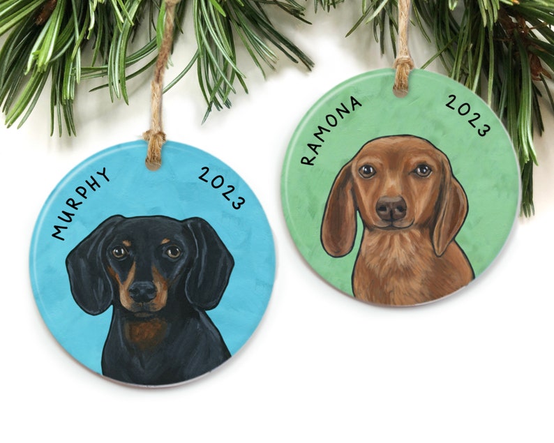 May include: Two round Christmas ornaments with dachshund dog portraits. The ornament on the left is blue with the name "MURPHY" and the year "2023". The ornament on the right is green with the name "RAMONA" and the year "2023".