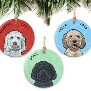 May include: Three round ceramic ornaments with dog portraits and names. The ornaments are red, blue, and green with the year 2023 printed on each. The dogs are a white doodle named Peaches, a golden doodle named Bella, and a black doodle named Benji.