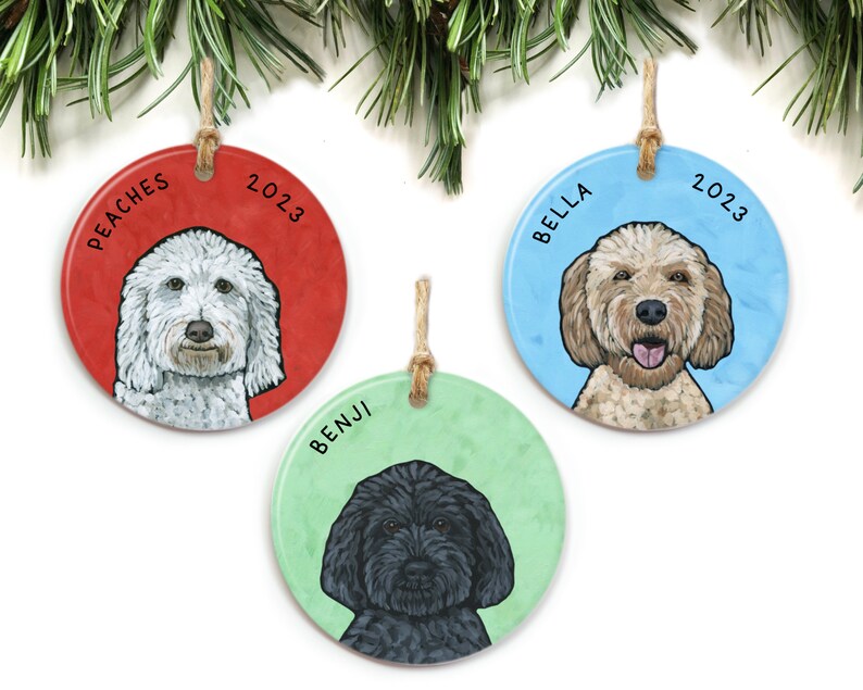 May include: Three round ceramic ornaments with dog portraits and names. The ornaments are red, blue, and green with the year 2023 printed on each. The dogs are a white doodle named Peaches, a golden doodle named Bella, and a black doodle named Benji.