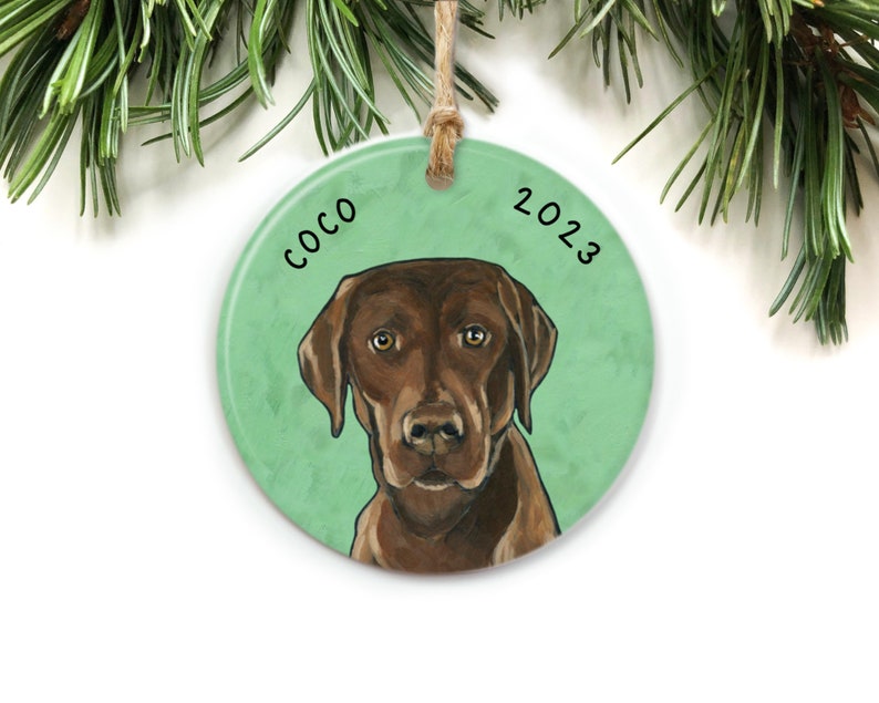 Puede incluir: Un adorno navide&ntilde;o redondo de cer&aacute;mica con fondo verde y un perro Labrador Retriever marr&oacute;n. El nombre del perro "Coco" y el a&ntilde;o "2023" est&aacute;n escritos en el adorno.