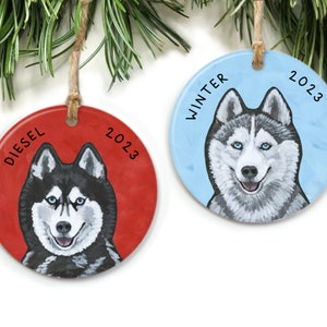 May include: Two round ceramic Christmas ornaments featuring a husky dog. One ornament is red with the name "Diesel" and the year "2023". The other ornament is blue with the word "Winter" and the year "2023".