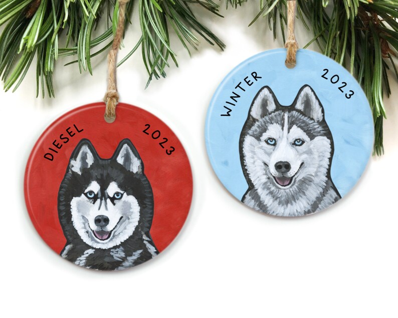 May include: Two round ceramic Christmas ornaments featuring a husky dog. One ornament is red with the name "Diesel" and the year "2023". The other ornament is blue with the word "Winter" and the year "2023".
