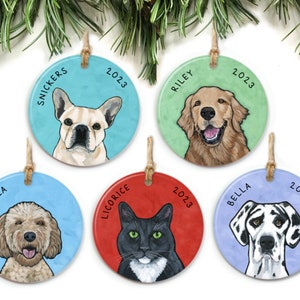 May include: Five round ceramic ornaments with painted portraits of pets. Each ornament has a blue, green, red, or purple background with the pet's name and the year 2023 written on it. The pets include a French Bulldog, a Golden Retriever, a Goldendoodle, a black and white cat, and a Great Dane.