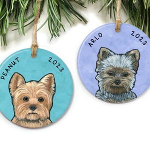 May include: Two round ceramic ornaments with a painted Yorkshire Terrier dog on each. The ornament on the left is blue with the text "PEANUT 2023". The ornament on the right is purple with the text "ARLO 2023".