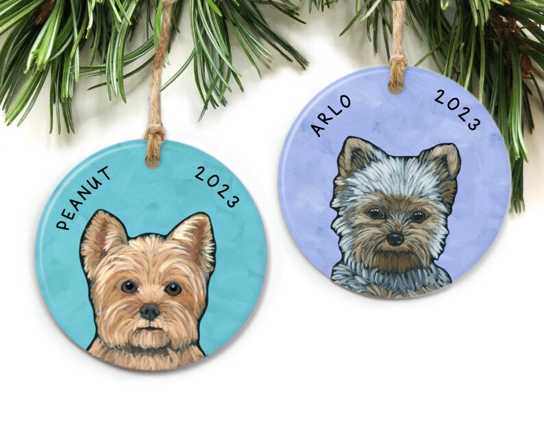 May include: Two round ceramic ornaments with a painted Yorkshire Terrier dog on each. The ornament on the left is blue with the text "PEANUT 2023". The ornament on the right is purple with the text "ARLO 2023".