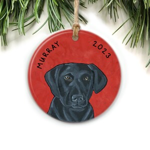Puede incluir: Un adorno navide&ntilde;o redondo y rojo con un retrato de un Labrador Retriever negro. El nombre del perro, "MURRAY", est&aacute; escrito en negro en el lado izquierdo del adorno. El a&ntilde;o "2023" est&aacute; escrito en negro en el lado derecho del adorno.
