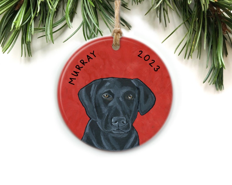 Puede incluir: Un adorno navide&ntilde;o redondo y rojo con un retrato de un Labrador Retriever negro. El nombre del perro, "MURRAY", est&aacute; escrito en negro en el lado izquierdo del adorno. El a&ntilde;o "2023" est&aacute; escrito en negro en el lado derecho del adorno.