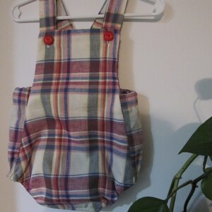 May include: A plaid baby onesie with red buttons and straps. The onesie is hanging on a white hanger against a white wall.