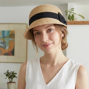 May include: A beige cloche hat with a black ribbon band and bow. The hat features a rounded crown and a gently sloping brim. The hat is made of a woven material, and the black ribbon provides a contrasting detail.