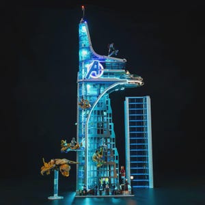 May include: A detailed model of the Avengers Tower, illuminated with blue lights. The building features the Avengers logo and several miniature vehicles and figures. The model is set against a dark background, highlighting the glowing details.