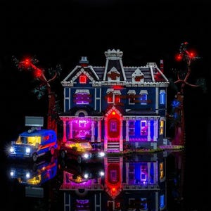 Può includere: Un modello dettagliato di una casa blu con finestre illuminate e una porta d'ingresso rossa. Due veicoli, un furgone blu e un'auto nera, sono parcheggiati di fronte. La scena è ambientata su uno sfondo nero, con illuminazione rossa e viola.
