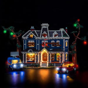 Könnte beinhalten: Ein blau-weißes Lego-Haus mit beleuchteten Fenstern und einer Veranda, flankiert von Bäumen mit farbigen Lichtern. Ein blauer Van und ein rotes Auto mit Scheinwerfern stehen vor dem Haus, vor schwarzem Hintergrund.