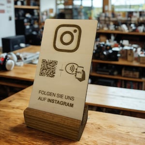 Könnte beinhalten: Ein hölzernes Instagram-Schild mit dem Instagram-Logo, einem QR-Code und dem Text "FOLGEN SIE UNS AUF INSTAGRAM". Das Schild wird auf einem Holzständer präsentiert, ideal für ein Unternehmen oder Geschäft.