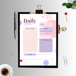 Puede incluir: Un portapapeles negro contiene un planificador diario con un diseño rosa y morado. El planificador incluye secciones para la fecha, el horario, la prioridad, las notas y las tareas de mañana. También se ven un bolígrafo negro, clips y una taza de café.