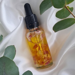 Op de afbeelding: Een heldere glazen fles met een zwarte druppelkap gevuld met gele vloeistof en bloemblaadjes. De fles staat op een witte satijnen ondergrond, met groene eucalyptusbladeren op de achtergrond. De fles lijkt essentiële olie te bevatten.